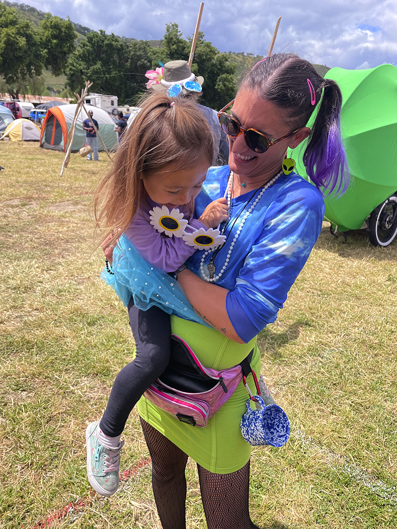 Festival nanny holding child at outdoor music festival with tents and colorful outfits